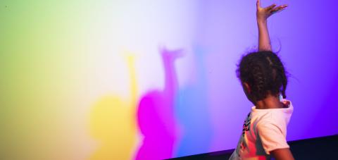 Lapsi käsi pystyssä, seinällä sateenkaaren värisiä varjoja.