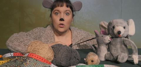 Hiireksi naamioitunut nainen, joka kertoo satua erilaisten pehmolelujen kera.