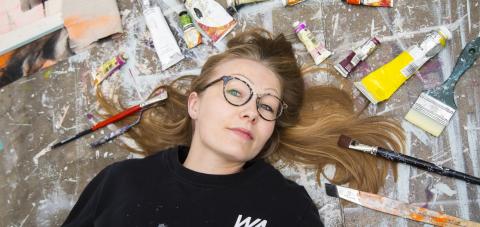 Silmälasipäinen nainen makaa lattialla. Hänen hiuksensa ovat pitkin lattiaa. Hiuksia kehystää lattialla olevat maalituubit ja pensselit. Lattialla on maaliroiskeita.