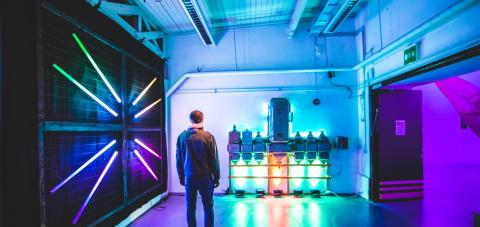 Takaapäin kuvattu mies on katselemassa valotaideteosta, jossa metallinen laite on valaistu vihreillä ja oranssilla valolla. Vasemmalla metalliverkkoseinällä on eri värisiä loisteputkia säteiksi aseteltuna. Huone on täynnä violettia, vihreää ja sinertävää valoa.