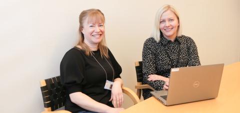 Kaksi henkilöä istuu pöydän ääressä tietokoneen kanssa ja katsovat hymyillen kameraan.