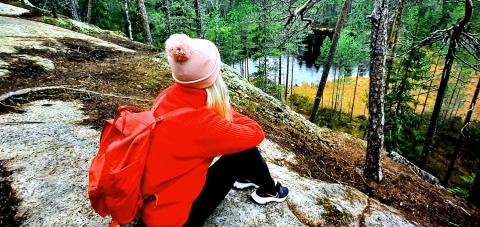 A girl in a red jacket with a rucksack on her back is sitting on a rock, looking out over a landscape with a forest and a small lake.