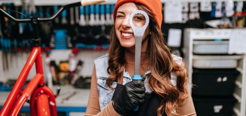 Pitkähiuksinen nuori henkilö kurkistaa toisella silmällä son kiintoavaimen reiän läpi. Taustalla pyöränkorjauspaja työkaluineen.