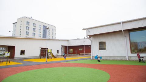 Hippoksen valkoisen puupäiväkodin pihalla näkyy värikäs pelialue.