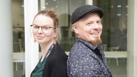 Kaksi ihmistä istuvat selät vastakkain ja hymyilevät kameralle.