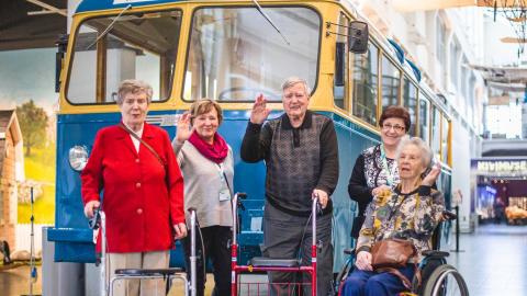 Viisi ikäihmistä seisoo museon käytävällä vanhan kaupunkibussin edessä. Osalla henkilöistä on rollaattori tai pyörätuoli. Kaksi heistä heilauttaa kättään kuvaajan suuntaan.