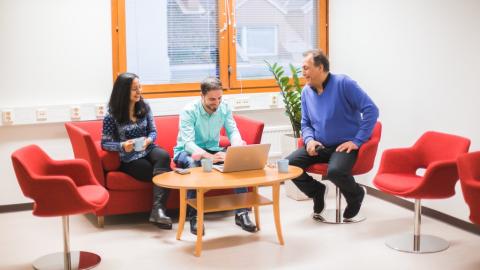 Yksi nainen ja kaksi miestä istuu toimistossa. Yksi heistä käyttää tietokonetta ja muut seuraavat työskentelyä