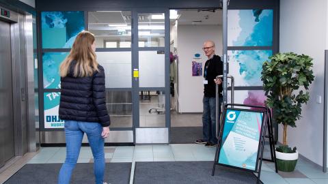 Työntekijä pitää ovea auki. Nuori henkilö on astumassa sisälle.