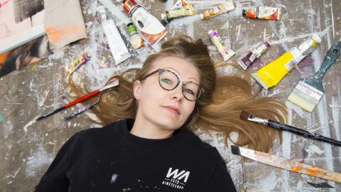 Silmälasipäinen nainen makaa lattialla. Hänen hiuksensa ovat pitkin lattiaa. Hiuksia kehystää lattialla olevat maalituubit ja pensselit. Lattialla on maaliroiskeita.