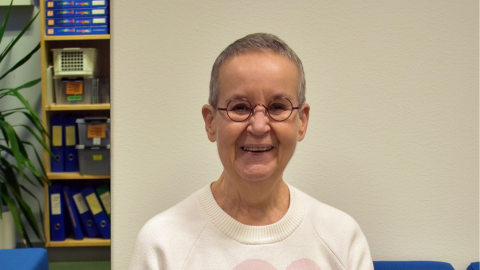 Henkilö istuu sohvalla ja katsoo kameraan. Taustalla peili, jossa näkyy mappeja.