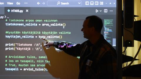 Henkilö seisoo älytaulun edessä. Taululla on Python-kielistä ohjelmointitekstiä.