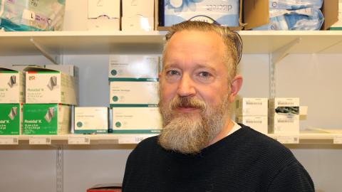 Parrakas mies hymyilee vienosti varastohyllyjen edessä. 