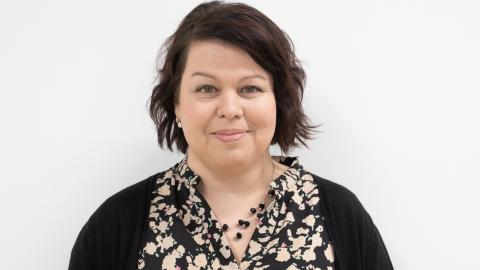 Hankkeen projektipäällikkö hymyilee kasvokuvassa. Tummat lyhyet hiukset, helmet kaulassa. 