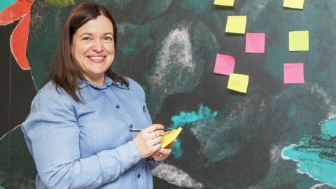 Kumpulainen kirjoittaa muistilapulle. Taustaseinälle on kiinnitetty kirkkaan keltaisia ja pinkkejä muistilappuja.