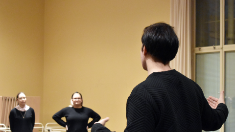 Henkilö seisoo salissa. Taka-alalla kaksi henkilöä kuuntelee ohjeistusta.