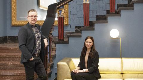 Heikki Aittala seisoo portaiden edessä ja Peppiina Inervo istuu sohvalla Tuulensuun palatsin aulassa, molemmat katsovat kameraa kohti hymyillen. 