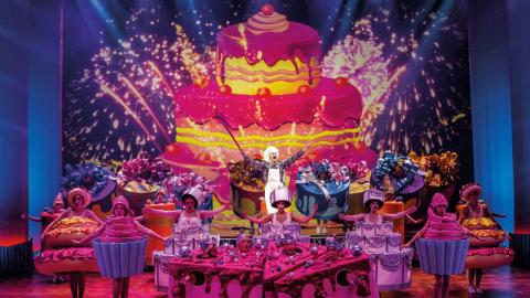 Näyttelijät tanssivat ja laulavat teatterin esiintymislavalla. Kaikki ovat pukeutuneet leivoksiksi tai kuppikakakuiksi. 