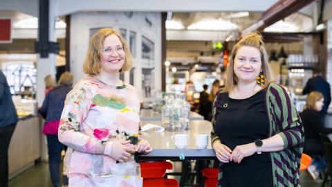 Kaksi henkilöä seisoo nojaten pöytään ja katsoo kameraan.