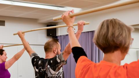 Kolme henkilöä keppijumppaa liikuntasalissa. Ylös nostetut kädet pitelevät puista keppiä.