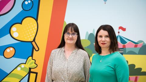 Tampereen kaupungin lapsiasiamiehet Tiia Heinäsuo ja Taru Kuosmanen seisovat rinnakkain. Heidän takanaan on värikäs, voimakkaasti kuvioitu seinä, jossa erottuu karkkeja ja sirkusteltta.
