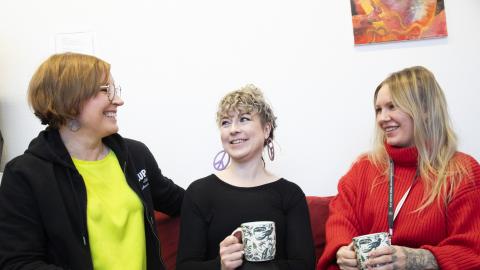 Kolme henkilöä istuu kahvikupit käsissään sohvalla.