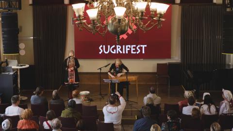 Suuren juhlasalin lavalla esiintyy laulaja ja kanteleen soittaja. Yleisö istuu penkeillä.