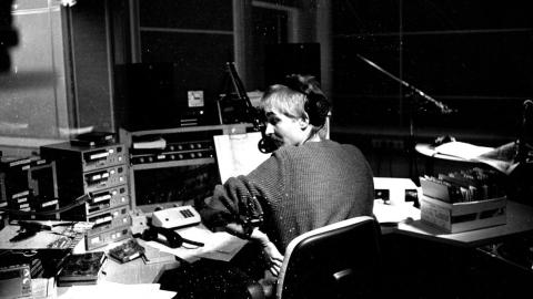 Mustavalkoisessa kuvassa mies istuu selkä kameraan päin kuulokkeet korvilla mikrofonin edessä.