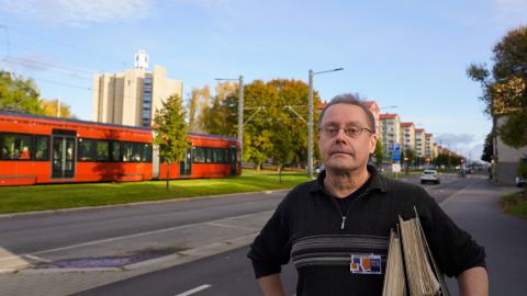 Henkilö seisoo kadulla. Hänen kainalossaan on arkistomappeja. Ratikka kulkee kadulla taustalla.