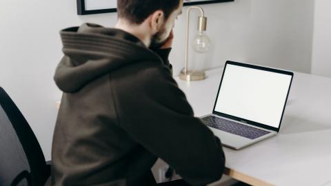 Tummaan huppariin pukeutunut nuori mies istuu pöydän ääressä ja katselee tietokoneen näyttöä. 