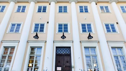 Vaalenkeltaisen ison rakennuksen julkisivua koristaa valkoiset pylväät. Julkisivulla on paljon ikkunoita.