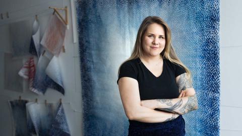 Mustahameinen, pitkähiuksinen nainen seisoo sinisävyisen seinälle ripustetun taideteoksen edessä. 