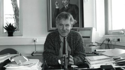 Henkilö istuu työhuoneessaan. Taustalla on suuri taulu ja pöydällä runsaasti papereita.