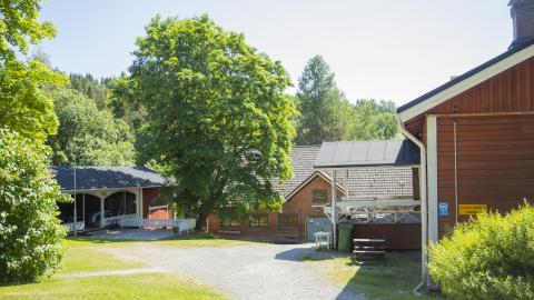 A court surrounded by trees. On the right an end of a red building, in the middle a red house, on the left a red house with an open porch and a terrace in front of it.