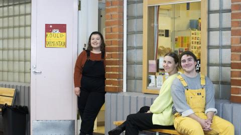 Vasemmalla seisoo kouluvalmentaja, joka nojaa seinään. Penkissä selät vastakkain istuu kaksi muuta kouluvalmentajaa.