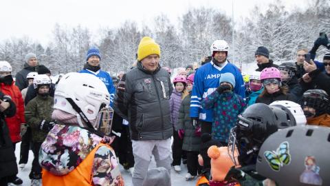 Iso joukko ihmisiä jäällä, suurin osa lapsia. Keskellä aikuinen mies keltainen pipo päässä, hänen molemmilla puolillaan seisoo mies. Useimmilla ihmisillä on päässään kypärä, joillakin pipo ja monilla on luistimet jalassa.