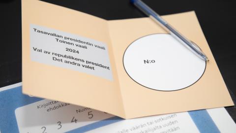 Tasolla ruskea äänestyslippu, jonka päällä kynä. Alla paperi, jossa numeromalleja.