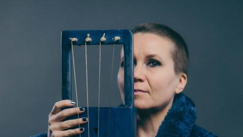 Lyhythiuksinen nainen pitelee siniseksi maalattua jouhikkoa puoliksi kasvojensa edessä. Naisella on sininen turkistakki.