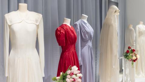 Hääpukuja mallinukkejen päällä näyttelytilassa. Puvut ovat pääasiassa valkoisia, mutta keskellä on sininen ja punainen hääpuku. Pukujen joukossa on kukkakimppuja.