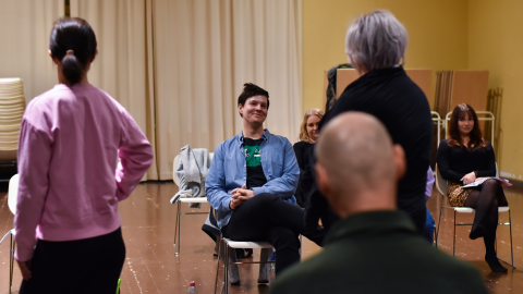 Opettaja istuu tuolilla. Hänen edessään on opiskelijoita esiintymässä.