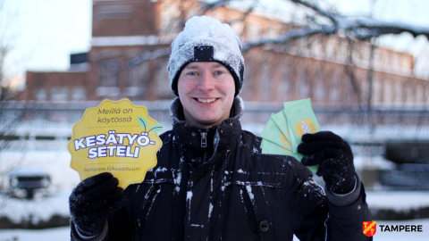 Kesätyösetelivastaava Petri Räsänen seisoo lumisena ulkona ja hymyilee. Hän pitelee käsissään isoa Kesätyösetelin tunnusta ja pieniä vihreitä Kesätyöseteleitä.