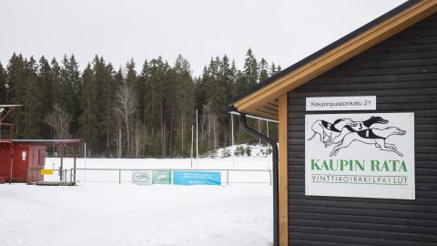 Etualalla vinttikoiraradalta ruskea rakennus, jossa vinttikoirilla kuvitettu kyltti. Itse rata ja toinen rakennus taustalla talvisessa maisemassa.