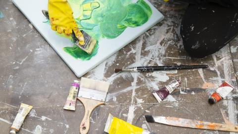 Lattialla maalituubeja, siveltimiä ja tekeillä oleva maalaus. Hanskaan verhottu käsi pitele sivellintä taulun päällä.