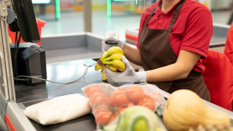 Kaupan kassahihnalla on erilaisia hedelmiä ja vihanneksia. Kassatyöntekijä, josta näkyvät vain kädet, on lukemassa niiden hintoja.