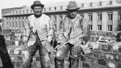 Kaksi hattupäistä muuraria istuu piipputiilien päällä.