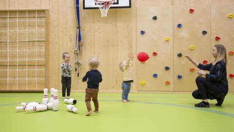 Kolme lasta ja päiväkodin työntekijä heittelevät palloja päiväkodin liikuntasalissa.