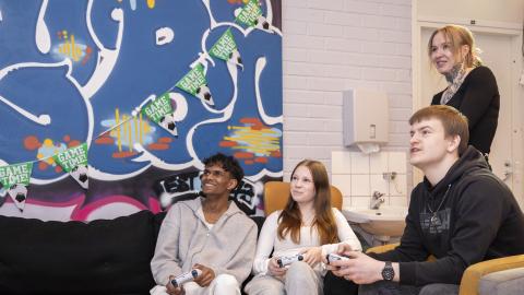 Neljä ihmistä huoneessa. Kolme istuu, yksi seisoo. Istuvilla on käsissään jokin laite. 