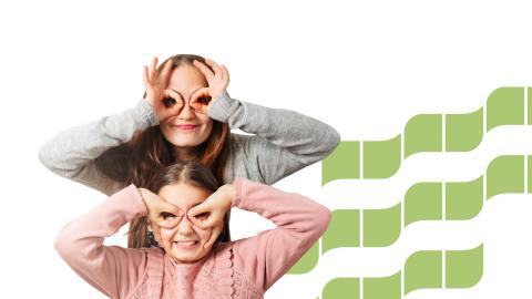 A mother stands behind her daughter, both smiling. They have made binoculars with their fingers, through which they are looking at the viewer.