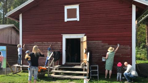 Vanhaa puurakennusta maalataan punaiseksi kesällä. Maalajat ovat aikuisia ja lapsia. Rakennuksen ovi on auki ja sisälle pääsee portaita.