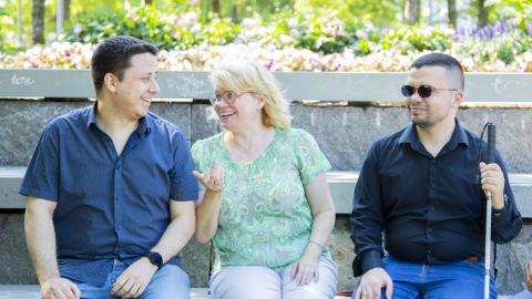 Kolme henkeä istuu rinnakkain puistossa keskustelemassa. Yhdellä heistä on näkövammaisen keppi ja mustat lasit.