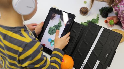 A preschool-aged child is taking pictures of fruits and vegetables with a tablet’s camera and using the Vegemi application to identify them.